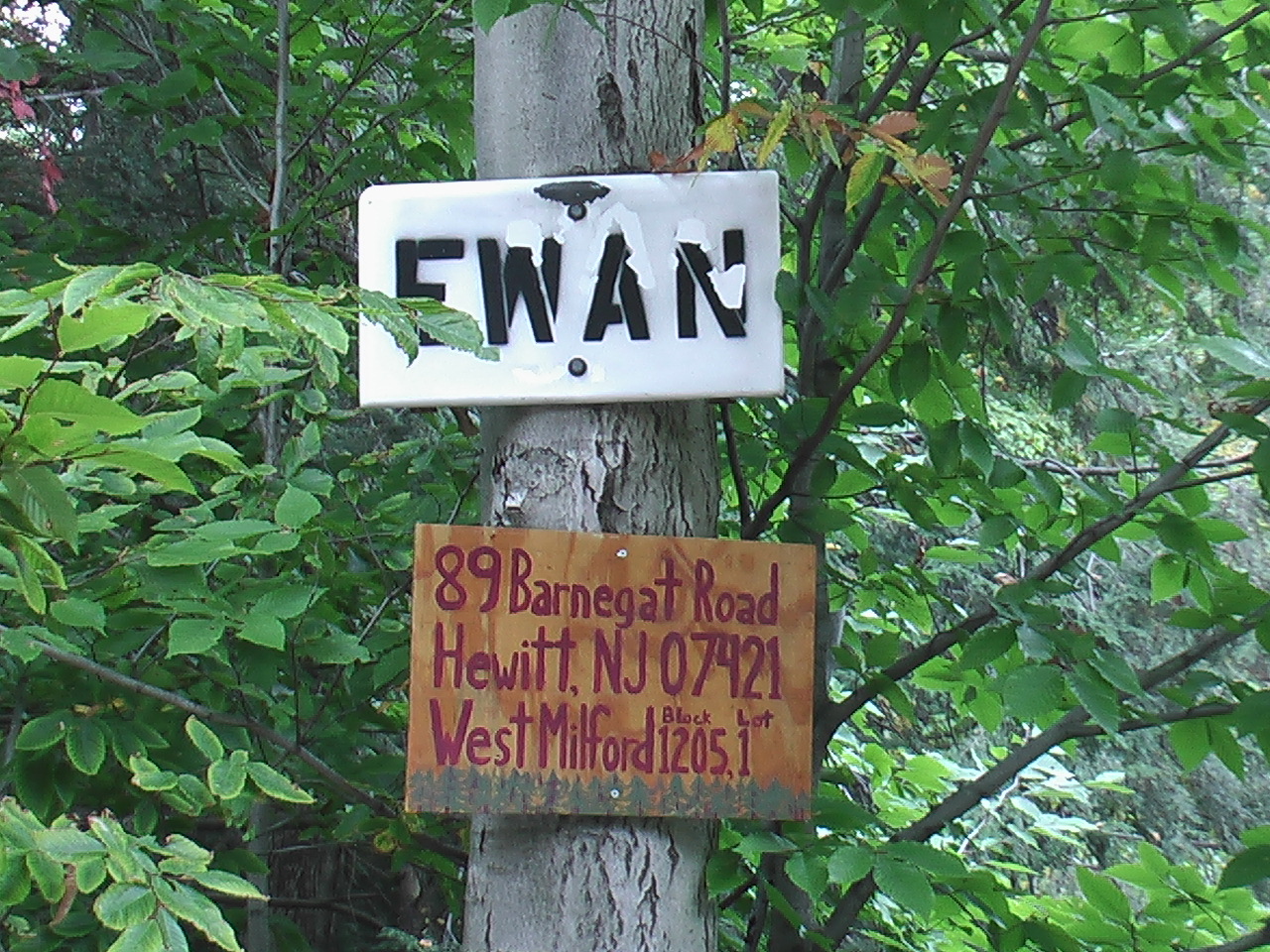 Image: Standing on southeast corner, there are two wood boards nailed on a tree, the upper board has capital letters EWAN that are colored black, and the lower board shows the address 89 Barnegat Road, West Milford (Hewitt), NJ 07421, Block 1205, Lot 1 and characters are colored burgundy.